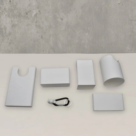 Five white BAGLETS tote bag accessories by Chatelet Manufacturing, including a phone case holder, key & wallet holder, carabiner key holder, bottle holder, and sunglasses holder, are arranged on a light surface with a gray wall in the background.