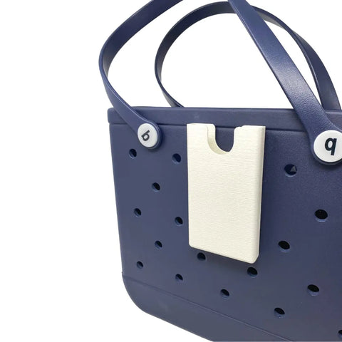 A navy blue rubber tote bag with round holes, two handles, and white b and q buttons features a white BOGLETS Phone Holder Charm Accessory by Chatelet Manufacturing clipped to the front to keep your phone handy.