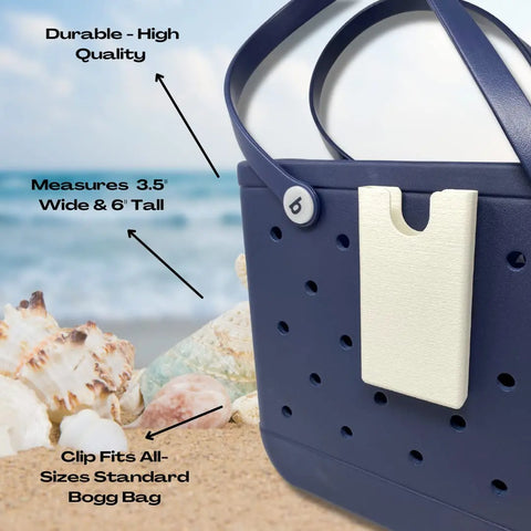 The BOGLETS Phone Holder Charm Accessory by Chatelet Manufacturing is shown clipped to a navy blue beach tote on sand with seashells. Text notes its durable quality, ideal 3.5 x 6 size, and universal clip for standard rubber beach tote bags. Made in the USA.