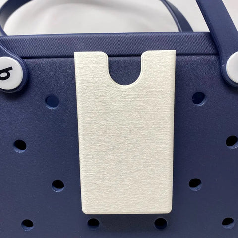Close-up of a navy blue tote bag with round holes and visible white letter buttons, featuring the BOGLETS Phone Holder Charm Accessory by Chatelet Manufacturing, compatible with Bogg Bags to keep your phone handy. Made in the USA.