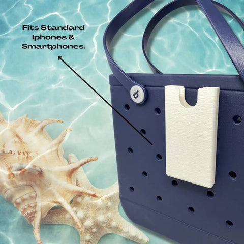 A BOGLETS Phone Holder Charm Accessory by Chatelet Manufacturing, attached to a dark blue rubber beach tote near seashells poolside. Text: Fits Standard iPhones & Smartphones, with an arrow to the phone holder. Made in the USA.