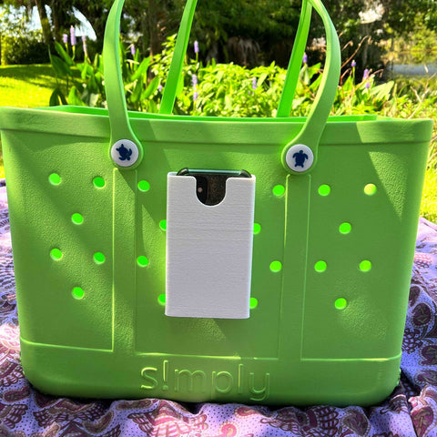 A large green tote bag with simply on the front rests outdoors. Attached is a BOGLETS phone holder charm accessory by Chatelet Manufacturing, keeping a smartphone handy while sunlight streams through the trees.