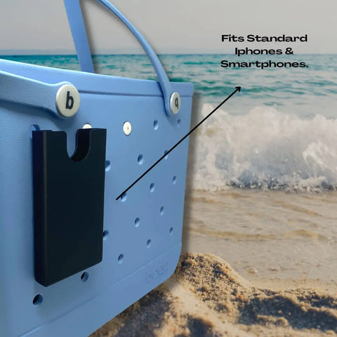 A BOGLETS Phone Holder Charm Accessory by Chatelet Manufacturing, attached to a blue beach tote bag on sand with the ocean behind. Text: Fits Standard iPhones & Smartphones, with an arrow pointing to the phone holder.