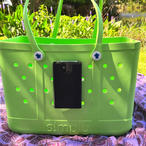 A bright green tote bag with simply embossed sits outdoors on a blanket, featuring a Chatelet Manufacturing BOGLETS phone holder charm accessory—compatible with Bogg Bags—attached to the front and lush greenery in the background.