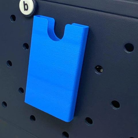 A blue rectangular BOGLETS phone holder charm accessory by Chatelet Manufacturing is attached to a black pegboard with round holes. A white peg labeled b appears at the top left.