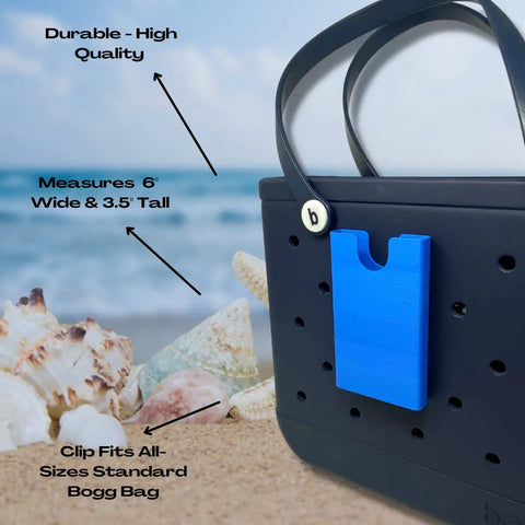 A blue BOGLETS phone holder charm by Chatelet Manufacturing clips to a black tote with holes, shown on sand near seashells. Arrows highlight: Durable & High Quality, 6x3.5”, and Fits All Standard Bogg Bags. Made in the USA.