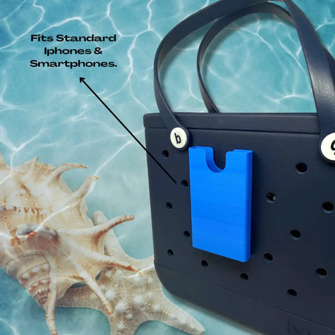 A black beach tote bag with a blue BOGLETS phone holder charm accessory by Chatelet Manufacturing, shown near seashells on a blue water-patterned background. Text reads: Fits Standard iPhones & Smartphones. Made in the USA.