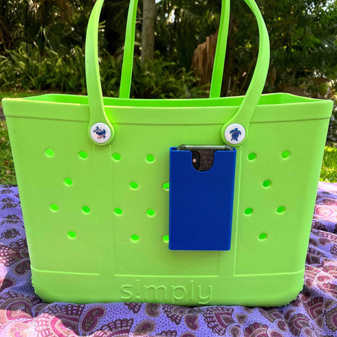 A green rubber beach tote with round handles and small holes sits outside. Attached is a blue BOGLETS Phone Holder Charm Accessory by Chatelet Manufacturing, holding a phone and featuring cartoon buttons near the handles.