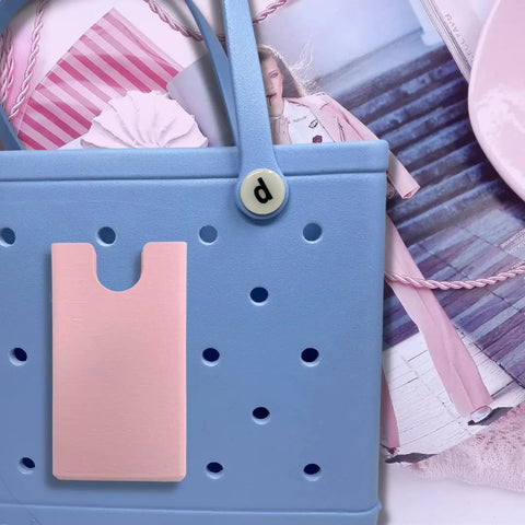 A pastel blue beach tote with a pink pocket and round “p” button from Chatelet Manufacturing sits on a fashion magazine. BOGLETS phone holder charm accessories, compatible with Bogg Bags, are scattered around. Made in the USA.