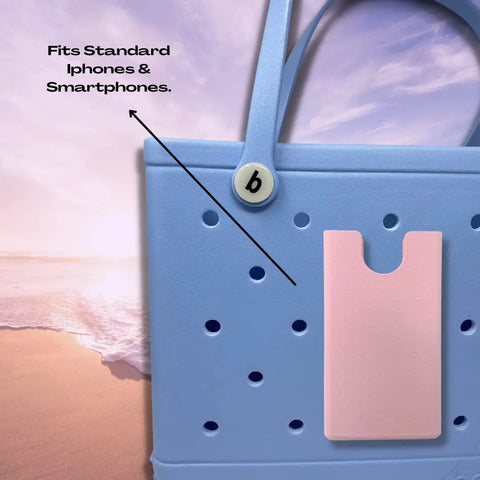 A blue tote bag with handles features a pink BOGLETS Phone Holder Charm Accessory by Chatelet Manufacturing, keeping your phone handy. Compatible with Bogg Bags and fits standard iPhones & smartphones. Made in the USA.