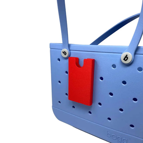 A light blue rubber beach tote bag with perforated holes, two blue handles, and a Chatelet Manufacturing BOGLETS Phone Holder Charm Accessory in red attached to the front to keep your phone handy.