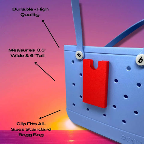 A BOGLETS Phone Holder Charm Accessory by Chatelet Manufacturing, compatible with all standard Bogg Bags, is shown on a blue beach tote with a red clip at sunset. Durable, 3.5 x 6, and made in the USA to keep your phone handy.