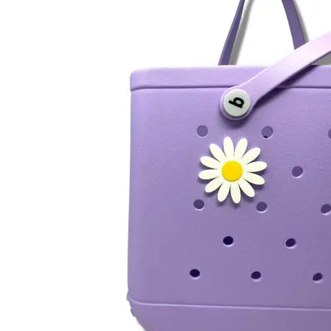 A light purple tote bag with round holes features a BOGLETS Flower Charm Accessory by Chatelet Manufacturing, perfect for customizing your Bogg Bag, and a snap button on the strap, shown against a white background.