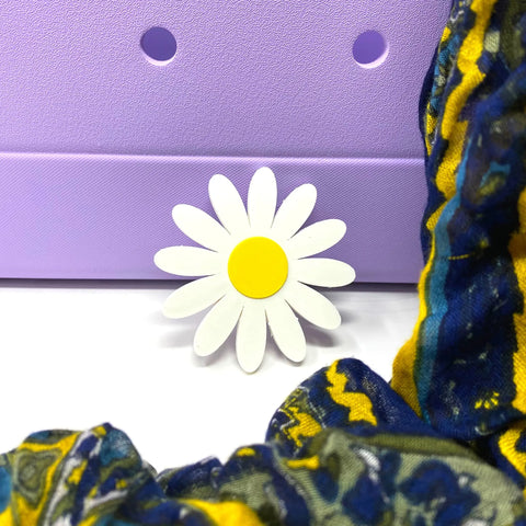 The BOGLETS Flower Charm Accessory by Chatelet Manufacturing, a cute decorative charm compatible with Bogg Bags, is displayed before a light purple textured surface and vibrant blue-yellow fabric, perfect for customizing your bag.