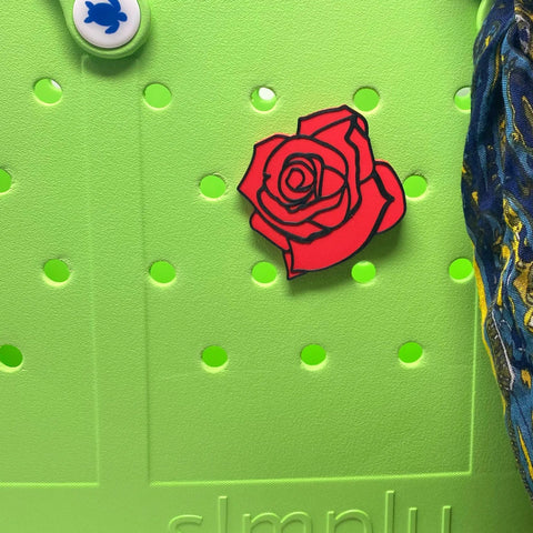 A BOGLETS Flower Charm Accessory by Chatelet Manufacturing, featuring a red rose pin and a flower bag charm, decorates a light green Bogg Bag with round holes. A blue and yellow-patterned fabric and part of the word simply are also visible.