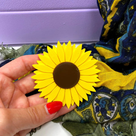 A hand with red-painted nails holds the BOGLETS Flower Charm Accessory by Chatelet Manufacturing—a bright yellow sunflower-shaped charm for Bogg Bags—shown against a colorful scarf and a light purple box.