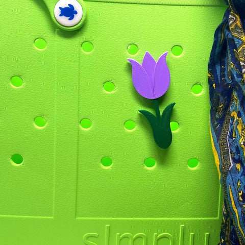 A close-up of a bright green Bogg Bag with round holes, decorated with a Chatelet Manufacturing BOGLETS Flower Charm accessory and a blue-patterned fabric draped on the right side.
