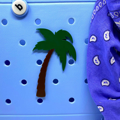 A BOGLETS Beach Charm Accessory by Chatelet Manufacturing is attached to a light blue pegboard, with a white b button and a purple bandana with white patterns nearby. Perfect for personalizing your Bogg Bag.