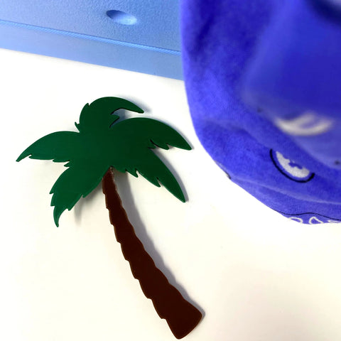 A flat, plastic palm tree with green leaves and a brown trunk lies on a white surface near a blue and purple object—BOGLETS by Chatelet Manufacturing, perfect decorative charms to personalize your Bogg Bag.