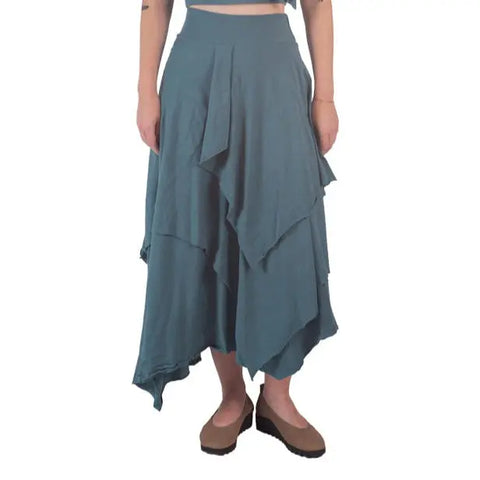 A person models the Spiritex Tri Fab Layered Skirt—a teal Bohemian piece made of organic cotton—paired with brown platform shoes. Only the lower body is shown against a white background.