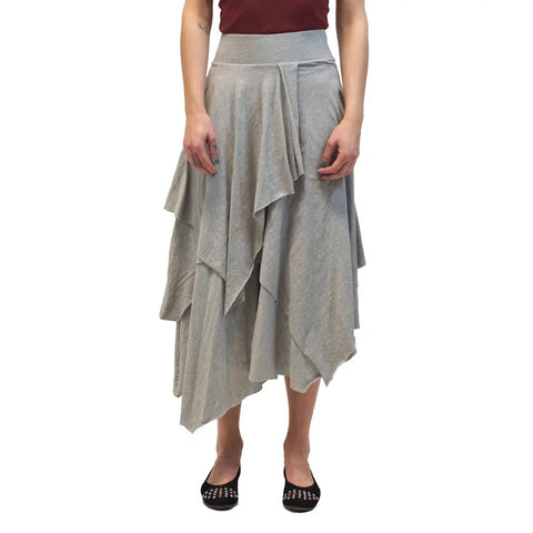 A person wears the Tri Fab Layered Skirt by Spiritex | Sustainable Apparel, a gray bohemian organic cotton piece, paired with a burgundy top and black studded flats, standing against a white background.