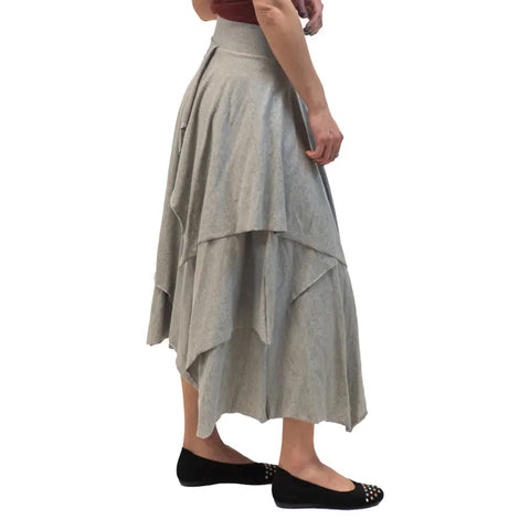 A person wearing the Tri Fab Layered Skirt by Spiritex | Sustainable Apparel in light gray, black flats with gold studs, and a short-sleeved top, stands against a white background.
