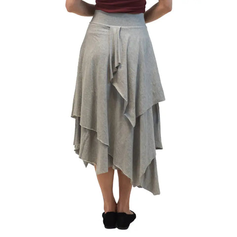 A person is shown from the back wearing black shoes and the Tri Fab Layered Skirt by Spiritex | Sustainable Apparel, a light gray organic cotton bohemian skirt with a soft drape and uneven hem.