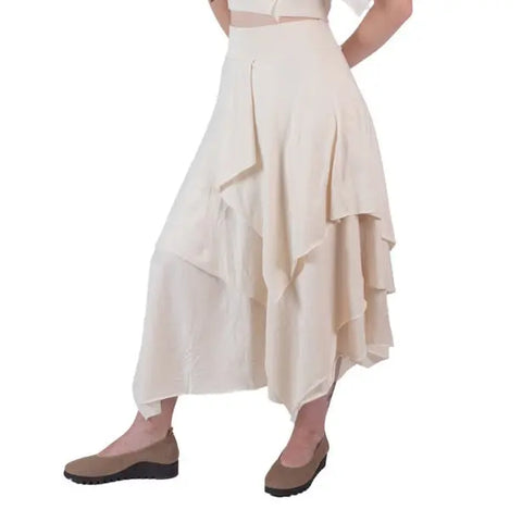 A person wearing the Spiritex Tri Fab Layered Skirt, a cream asymmetrical organic cotton piece, with brown flats. Shown from the waist down against a white background.