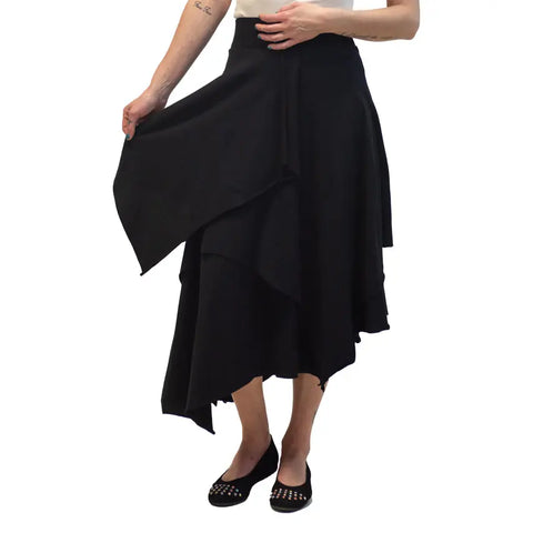 A person stands against a white background holding out one side of the black Spiritex | Sustainable Apparel Tri Fab Layered Skirt, paired with black studded flat shoes.