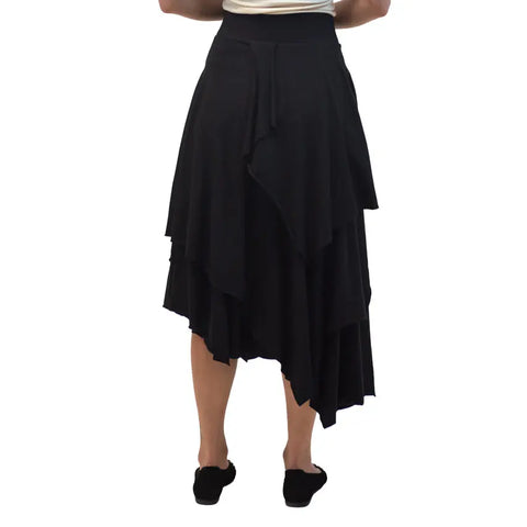 A person faces away from the camera against a white background, wearing the Spiritex | Sustainable Apparel Tri Fab Layered Skirt in black and matching black shoes.