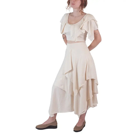 A woman wearing the Spiritex | Sustainable Apparel Boho Top in cream with ruffles and a matching organic cotton skirt stands against a white background, hands behind her back, and completes the look with brown flats.