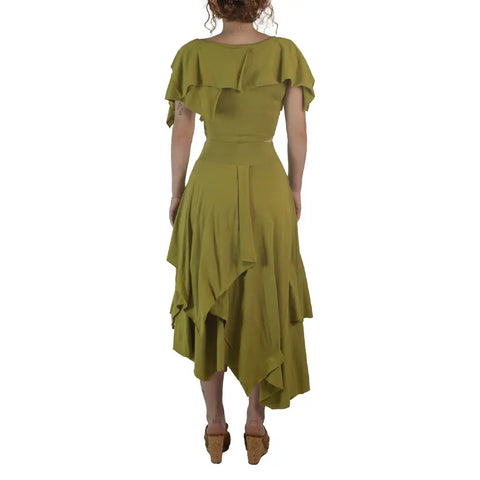 A woman faces away, wearing the Boho Top by Spiritex | Sustainable Apparel—a light olive green, off-shoulder organic cotton piece with layered asymmetrical ruffles. She pairs it with wedge sandals against a white background.
