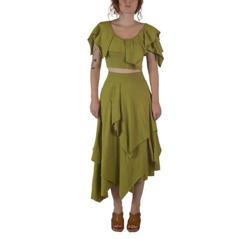 A woman faces forward in a green Boho Top by Spiritex | Sustainable Apparel with an asymmetrical, layered organic cotton skirt. She has curly hair, brown sandals, and her arms are relaxed at her sides.
