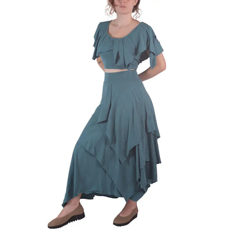 A woman in a teal Boho Top by Spiritex | Sustainable Apparel and a layered, asymmetrical organic cotton skirt with brown shoes stands against a white background.