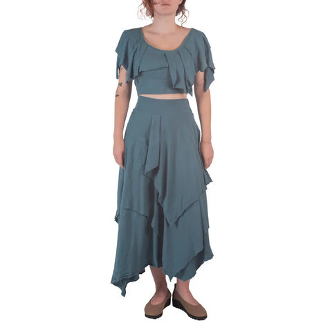 A woman models the Spiritex | Sustainable Apparel Boho Top in teal, paired with a matching asymmetrical skirt made from organic cotton, standing before a white background.