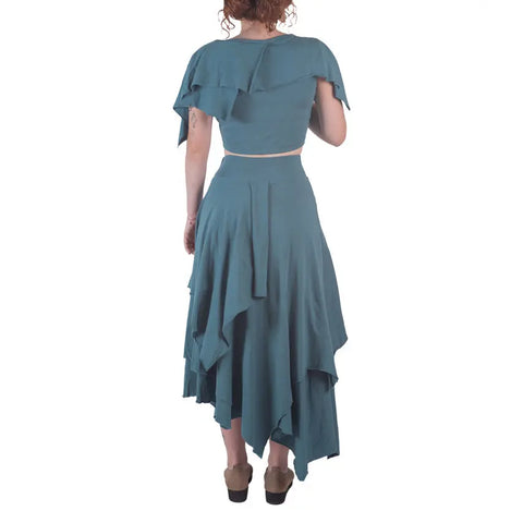 A woman with curly hair, seen from behind, wears a teal Boho Top by Spiritex | Sustainable Apparel and an asymmetrical skirt with brown shoes, standing against a white background.