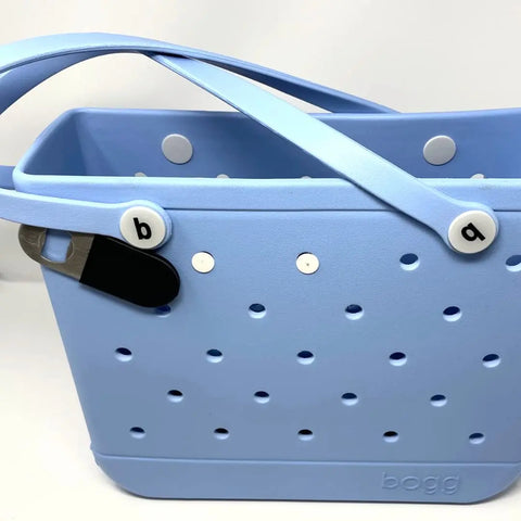 A light blue Bogg Bags tote with round holes and two crossed handles features the Chatelet Manufacturing BogLets Bottle Opener Charm accessory, easily attached for convenience. The bag has “bogg” embossed at the bottom and white b&q tags.
