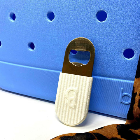 A BOGLETS PREMIUM Bottle Opener New 2023 Edition Accessory Charm by Chatelet Manufacturing, featuring a white textured handle, leans on a blue Bogg Bag with part of a leopard print fabric tote visible at the images edge.