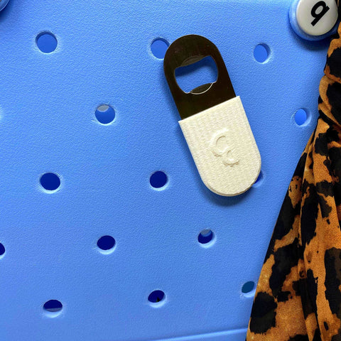 A BOGLETS PREMIUM Bottle Opener (White) from Chatelet Manufacturing is attached to a blue perforated tote bag, serving as a decorative accessory. A section of leopard-print fabric appears on the right side.