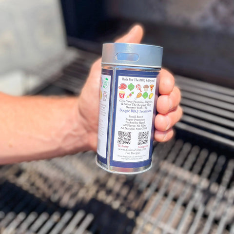 A hand holds a Gustus Vitae Open Fire BBQ Collection - 3 Pack container by an open grill, clearly showing its artisanal BBQ seasonings and QR code on the label.