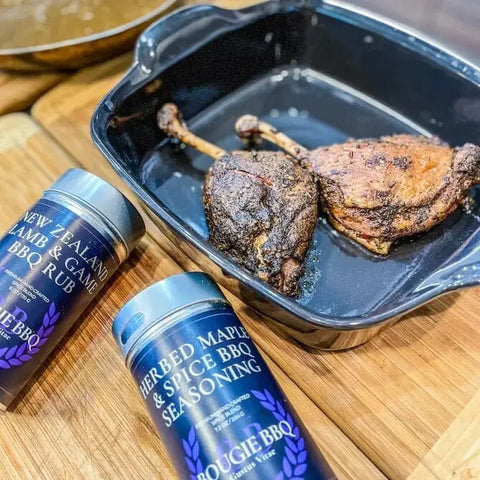 A black baking dish with two cooked duck legs sits on a wooden surface. Nearby are two Gustus Vitae Open Fire BBQ Collection seasonings from the 3 Pack: Lamb & Game BBQ Rub and Herbed Maple & Spice BBQ Seasoning.