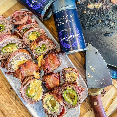 A platter of bacon-wrapped, cheese-stuffed jalapeño rolls sits by a cleaver and the Open Fire BBQ Collection - 3 Pack from Gustus Vitae, spotlighting their small batch BBQ rubs. In the background is a used baking tray with burnt bits.
