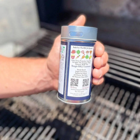 Someone holds a Gustus Vitae Complete Bougie BBQ Collection - Sweet 16 spice container by an open grill. The label shows colorful icons, branding, text, and a QR code, all clearly lit by bright outdoor light.