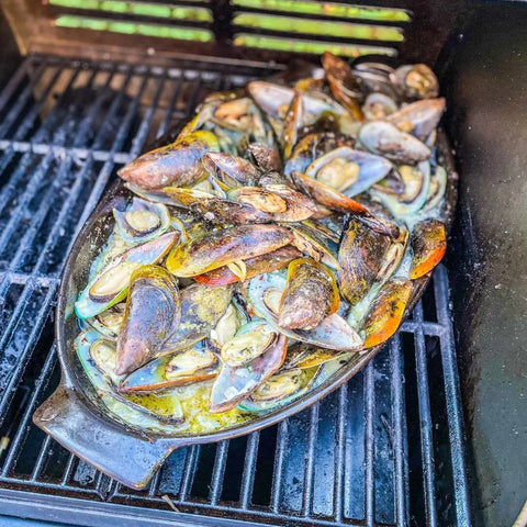 A ceramic dish of mussels in creamy sauce grills to perfection, their open shells revealing tender seafood, all enhanced by Gustus Vitae’s Complete Bougie BBQ Collection - Sweet 16 artisanal spice blends.
