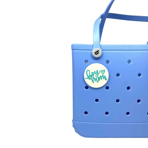 A partial view of a light blue tote features the Chatelet Manufacturing FRESHe BOGLETS Boy Mom charm (teal and white) attached to the handle—a trendy decorative accessory compatible with Bogg Bags, shown against a white background.