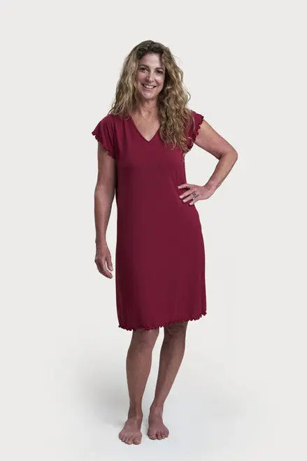 A woman with long, curly hair smiles while standing barefoot in a Goodnighties Flutter Sleeve Knee Length Gown, featuring 37.5 thermoregulating technology, against a plain white background.