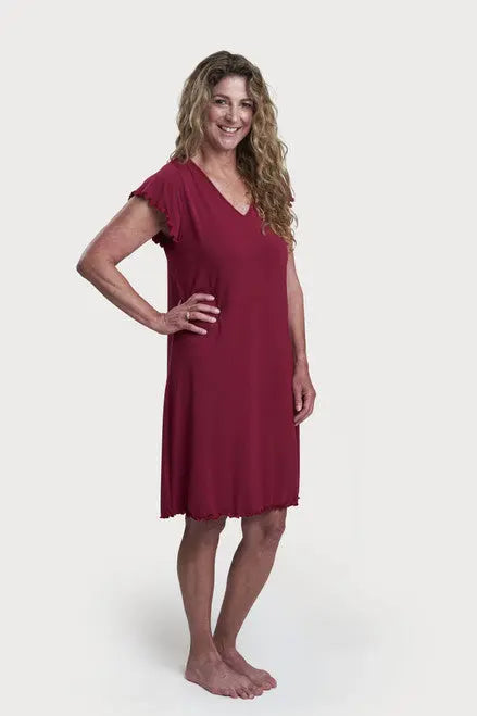 A woman with long, wavy hair stands barefoot and smiling against a plain white background, wearing the Goodnighties Flutter Sleeve Knee Length Gown with thermoregulating short sleeves.