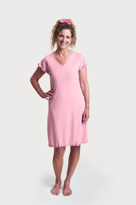 A smiling woman with curly hair wears the Goodnighties Flutter Sleeve Knee Length Gown with 37.5 technology and a matching pink scrunchie, standing barefoot against a plain white background.