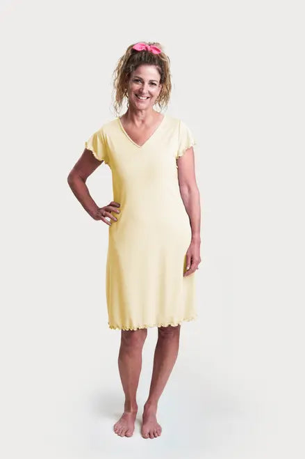 A smiling woman with curly hair in a pink scrunchie stands barefoot, modeling the Goodnighties Flutter Sleeve Knee Length Gown—yellow, short-sleeve, V-neck with scalloped edges—against a plain white background.