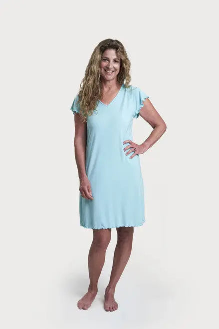 A woman with long wavy hair smiles barefoot on a white background, wearing the Goodnighties Flutter Sleeve Knee Length Gown—a light blue V-neck nightgown with scalloped edges and flutter sleeves. One hand is on her hip.